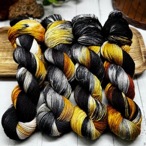 Puede incluir: Un ovillo de hilo teñido a mano en negro, dorado y blanco. El hilo está dispuesto en un patrón circular, mostrando las variaciones de color.