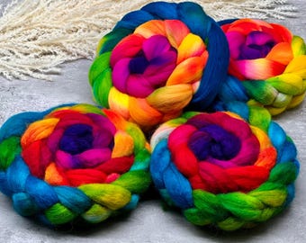 May include: Four circular braids of multi-colored wool roving featuring a vibrant gradient of blue, green, yellow, orange, red, and purple fibers. Each thick, soft coil is tightly wound into a round shape, showcasing a spectrum of colors.