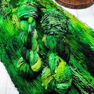 Könnte beinhalten: Zwei Stränge grüne Wolle mit schwarzen und gelben Akzenten liegen auf einem grünen Strickstoff. Das Garn ist eine Mischung aus hellen und dunklen Grüntönen, wodurch ein strukturiertes Aussehen entsteht. Der Hintergrund ist eine weiße Holzoberfläche.