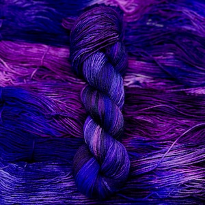 May include: A skein of purple and blue yarn with a gradient of color. The yarn is twisted into a braid and is lying on a background of similar yarn.