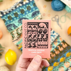 Könnte beinhalten: Ein rosa Kartenspiel mit dem Titel "Tropical Doodle Card Deck" mit schwarzen Illustrationen tropischer Elemente. Das Deck wird in einer Hand gehalten, mit bunter Wolle, Zitronen und Strickzubehör im Hintergrund.