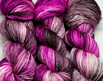 May include: Three skeins of yarn in shades of purple, white and black. The yarn is hand-dyed and has a variegated colour pattern.