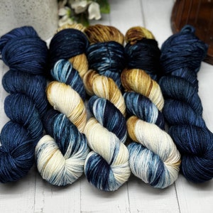 Op de afbeelding: Vijf bollen garen in tinten van blauw, crème en bruin. Het garen is waaiervormig gerangschikt op een wit houten oppervlak.