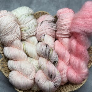 Könnte beinhalten: Ein Korb gefüllt mit rosa und weißen Garnknäueln. Das Garn ist weich und flauschig, perfekt zum Stricken oder Häkeln.