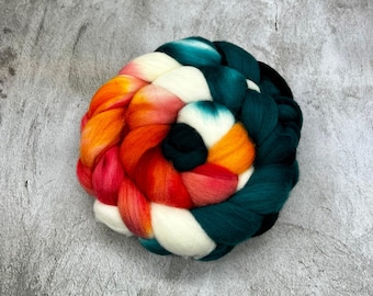 May include: A thick, braided coil of roving wool fiber featuring a vibrant color gradient. The wool transitions from deep teal and dark green into bright orange, red, and creamy white, arranged in a circular, spiraled pattern.