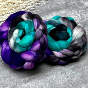 Può includere: Due spessi fasci intrecciati di lana cardata. Il fascio a sinistra presenta vivaci tonalità viola, turchese, nero e bianco. Il fascio a destra mostra una sfumatura di turchese, grigio scuro, nero e grigio chiaro, dall'aspetto morbido e testurizzato.