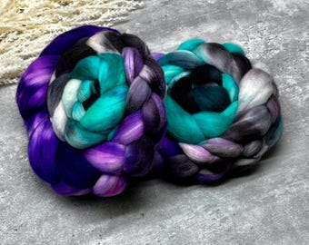 May include: Two thick, braided bundles of roving wool fiber. The left bundle features vibrant purple, teal, black, and white tones. The right bundle displays a gradient of teal, dark gray, black, and light gray, showcasing a soft, textured appearance.