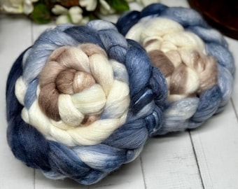 Può includere: Due spesse matasse intrecciate di lana cardata con una palette di colori sfumati in blu navy intenso, blu ardesia, crema e beige tenue. Le fibre presentano una consistenza liscia e lucente, avvolte in forme circolari floreali.