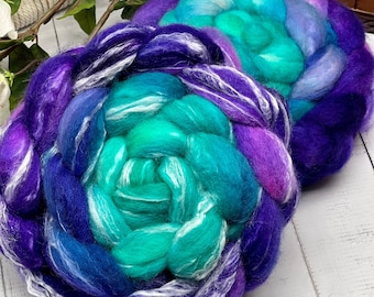 Puede incluir: Dos gruesas trenzas de lana cardada con una vibrante paleta de colores en degradado. Las fibras transicionan desde un violeta profundo y azul real en los bordes exteriores hacia un turquesa y verde azulado saturado en el centro.