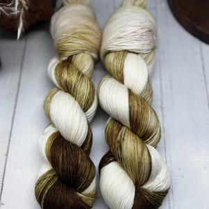 Puede incluir: Dos madejas de hilo teñido a mano en una paleta de colores naturales. El hilo es una mezcla de blanco, marrón y verde oliva, creando un efecto degradado. Las madejas están torcidas y listas para tejer o hacer ganchillo. Fondo blanco.