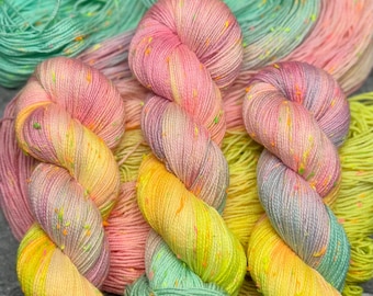 May include: Vibrant hand-dyed yarn skeins in pink, yellow, green, and purple. The yarn has a speckled appearance with small orange and yellow flecks. The skeins are arranged in a visually appealing display, ideal for knitting or crochet projects.