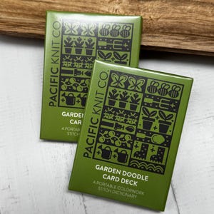 Könnte beinhalten: Zwei grüne Garden Doodle Kartenspiele mit schwarzen Illustrationen von Gartengeräten, Pflanzen und Insekten. Der Text "PACIFIC KNIT CO." und "GARDEN DOODLE CARD DECK" sind auf die Karten gedruckt. Die Karten liegen auf einer weißen Oberfläche.