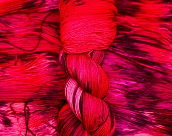 May include: A skein of red and black yarn with a variegated pattern. The yarn is twisted into a knot and is lying on a background of similar yarn.