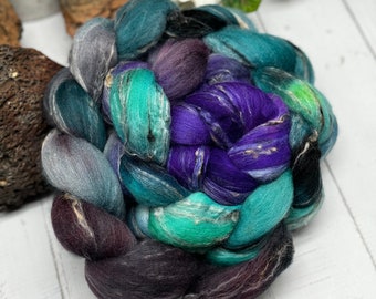 May include: A vibrant braid of hand-dyed fiber in shades of teal, purple, and gray. The fiber is arranged in a circular pattern, showcasing the blend of colors and textures. Ideal for spinning, felting, or other fiber art projects.