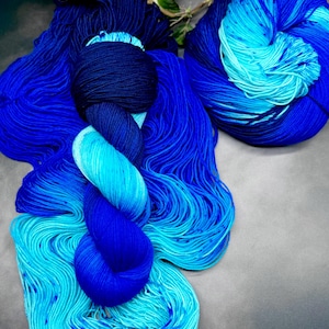 Op de afbeelding: Verschillende strengen blauw garen, variërend van diep marineblauw tot helder turquoise. Het garen is zo gerangschikt dat de kleurverlopen worden benadrukt. Op de achtergrond een paar takjes met witte bloemen en groene bladeren.