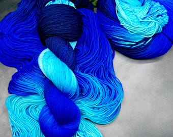 May include: A collection of blue yarn skeins in various shades, from deep navy to vibrant turquoise. The yarn is arranged to showcase the color gradients, with a few sprigs of white flowers and green leaves in the background.