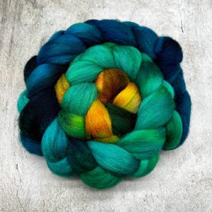 Può includere: Una spessa treccia di lana cardata non filata con una palette di colori sfumati. Gli strati esterni passano dal blu marino profondo al verde acqua, mentre il centro mostra tonalità vivaci di verde smeraldo, giallo brillante e arancione.