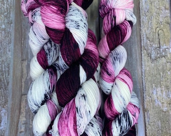 May include: Two skeins of yarn in a variegated pattern of white, black, and pink. The yarn is hand-dyed and has a speckled effect.