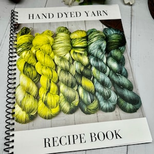 以下が含まれることがあります： 「HAND DYED YARN RECIPE BOOK」というタイトルのスパイラルバインディングされたレシピブック。表紙には、緑と黄色の色合いの手染め糸の糸巻きがいくつか写っています。