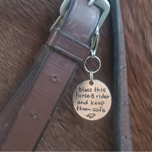 Może przedstawiać: Srebrny charm z tekstem "Bless this horse & rider and keep them safe" przymocowany do brązowego skórzanego haltera dla koni.
