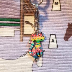 May include: A colorful cat charm with rainbow stripes and floral details hangs from a silver zipper pull. The charm is attached to a zipper on a purple and white fabric bag with a horse design.