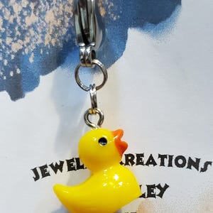 May include: A yellow rubber ducky charm with a silver clip. The charm is attached to a silver chain. The charm is labelled "JEWEL CREATIONS CLIP CHARMS".