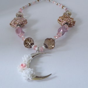 Op de afbeelding: Een delicate ketting met een halve maan hanger versierd met witte bloemen en roze accenten. De ketting heeft roze, witte en bruine kralen, waaronder een grote kristallen kraal.