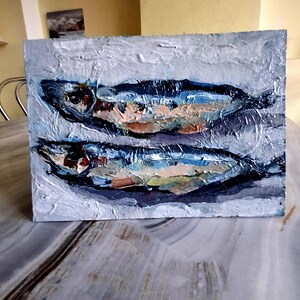 May include: An oil painting of two fish, rendered in a textured style. The fish are primarily blue, with accents of orange, white, and green. The background is a muted gray, creating a contrast that highlights the fish.