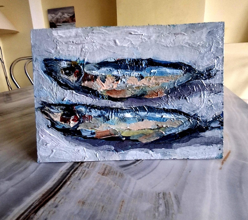 May include: An oil painting of two fish, rendered in a textured style. The fish are primarily blue, with accents of orange, white, and green. The background is a muted gray, creating a contrast that highlights the fish.