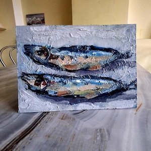 May include: Oil painting of two fish, rendered with thick brushstrokes. The fish are primarily blue, with accents of brown, beige, and white. The background is a textured, light gray. The artwork is on a rectangular canvas.