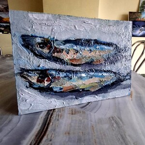 May include: An oil painting on canvas depicts two fish in shades of blue, brown, and white. The artwork has a textured, impasto style, with visible brushstrokes. The background is a light gray, and the painting is displayed on a table.