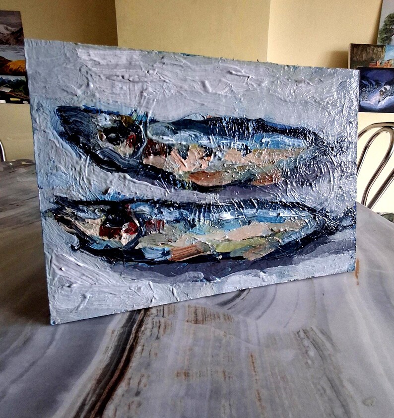 May include: An oil painting on canvas depicts two fish in shades of blue, brown, and white. The artwork has a textured, impasto style, with visible brushstrokes. The background is a light gray, and the painting is displayed on a table.