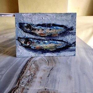May include: An oil painting on a square canvas depicts two fish in shades of blue, brown, and white. The textured brushstrokes create a sense of depth and movement. The artwork is set against a light gray background.