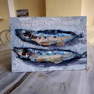 May include: An oil painting on a rectangular canvas depicts two fish. The fish are painted with blue, brown, and white brushstrokes, set against a textured, light gray background. The artwork has a rustic, impressionistic style.