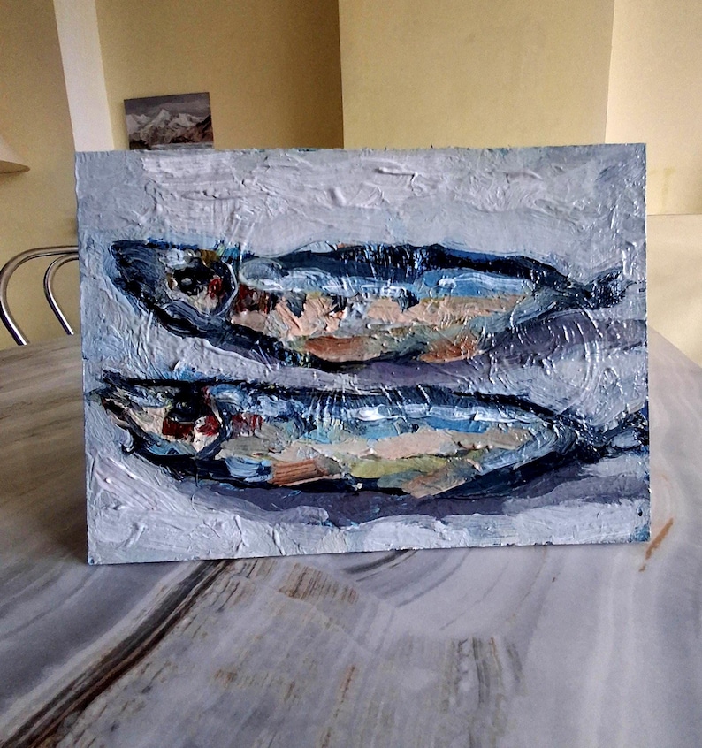 May include: An oil painting on a rectangular canvas depicts two fish. The fish are painted with blue, brown, and white brushstrokes, set against a textured, light gray background. The artwork has a rustic, impressionistic style.