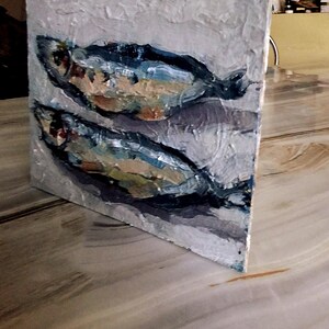 May include: Oil painting on canvas depicting two fish. The fish are painted in shades of blue, white, and brown against a textured, light gray background. The artwork is displayed on a white-edged square canvas.