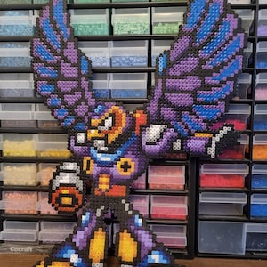 Può includere: Una figura pixel art di un personaggio con grandi ali, creata con perline di plastica colorate. La figura presenta una combinazione di colori viola, blu, nero e giallo. Il personaggio tiene in mano un oggetto rotondo.