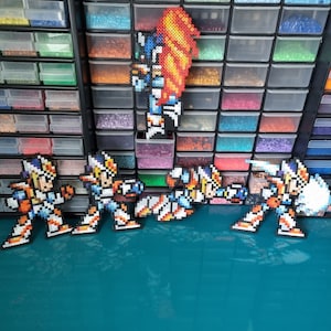 Könnte beinhalten: Vier Pixel-Art-Figuren eines Robotercharakters in den Farben Blau, Weiß und Orange. Die Figuren bestehen aus kleinen Plastikperlen und sind auf einer türkisfarbenen Oberfläche angeordnet. Eine fünfte Figur, ein Roboter mit einem feurigen orangefarbenen Umhang, befindet sich im Hintergrund.