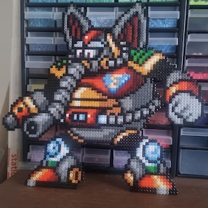 May include: A large, pixelated figure of a robot, created with colorful plastic beads. The robot has a red chest, gray arms and legs, and a blue gem on its chest. The robot's head is red and black, with pointed ears. The figure is standing on a brown surface.