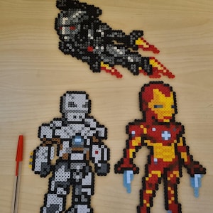 Può includere: Tre modelli di Iron Man realizzati con perline di plastica colorate. Uno è un jet nero e rosso, e gli altri due sono figure in piedi in rosso e oro, e argento e grigio. Una penna rossa è a sinistra.
