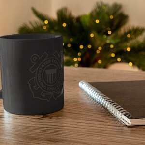 May include: Black ceramic coffee mug with a white graphic of the United States Coast Guard emblem. The emblem features an anchor, a shield with the American flag, and the words "United States Coast Guard 1790".
