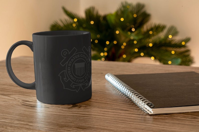 May include: Black ceramic coffee mug with a white graphic of the United States Coast Guard emblem. The emblem features an anchor, a shield with the American flag, and the words "United States Coast Guard 1790".