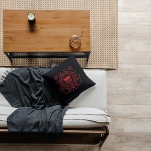 May include: A black throw pillow featuring a red United States Coast Guard emblem. The pillow is placed on a white sofa with a grey and black throw blanket. The sofa is positioned in front of a wooden coffee table with a candle and a vase.