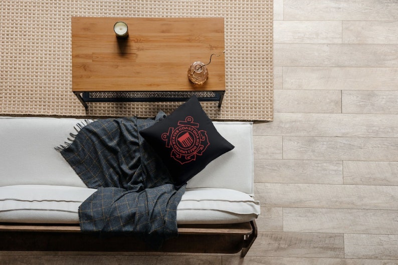 May include: A black throw pillow featuring a red United States Coast Guard emblem. The pillow is placed on a white sofa with a grey and black throw blanket. The sofa is positioned in front of a wooden coffee table with a candle and a vase.