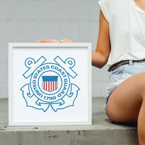 May include: A white framed print of the United States Coast Guard emblem. The emblem is blue and white and features an anchor, a shield with the American flag, and the words "United States Coast Guard 1790".