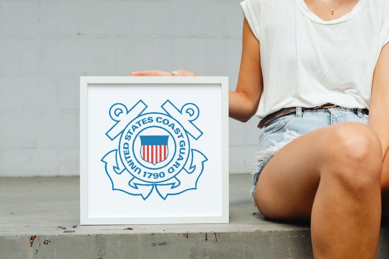 May include: A white framed print of the United States Coast Guard emblem. The emblem is blue and white and features an anchor, a shield with the American flag, and the words "United States Coast Guard 1790".
