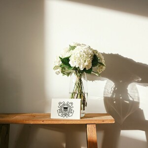 May include: A white hydrangea bouquet in a clear glass vase sits on a wooden table with a white card featuring the United States Coast Guard emblem.