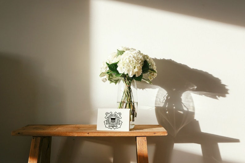 May include: A white hydrangea bouquet in a clear glass vase sits on a wooden table with a white card featuring the United States Coast Guard emblem.