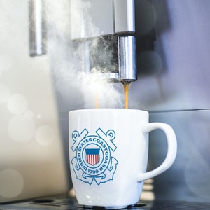 May include: A white coffee mug with the United States Coast Guard emblem printed on it. The mug is being filled with coffee from a silver coffee machine.