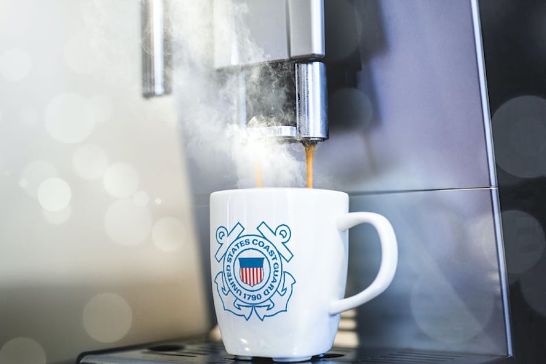 May include: A white coffee mug with the United States Coast Guard emblem printed on it. The mug is being filled with coffee from a silver coffee machine.